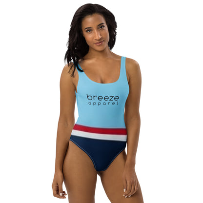 Woman wearing one-piece swimsuit with light-blue top and 'breeze apparel' in black across the chest, and a red stripe, then a white stripe, followed by a navy-blue bottom part.