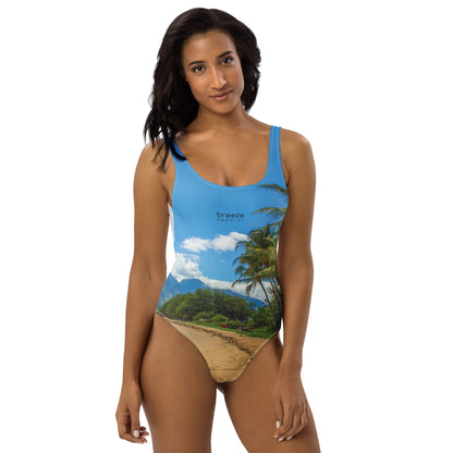 Woman wearing one-piece swimsuit with a coastline depicted on it. There is a clear sky, palm trees, mountains, and a beach depicted.