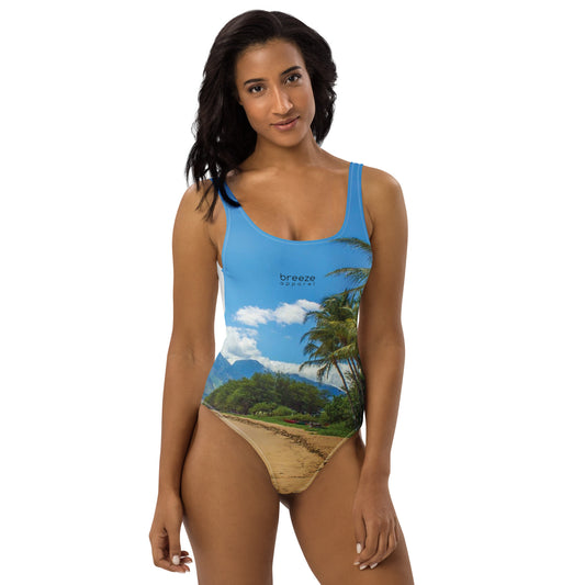 Woman wearing one-piece swimsuit with a coastline depicted on it. There is a clear sky, palm trees, mountains, and a beach depicted.