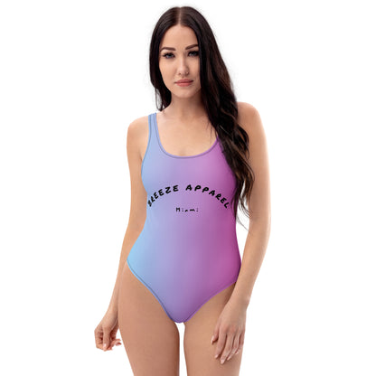 Woman wearing one-piece swimsuit with a light-blue-to-pink design and a black 'Breeze Apparel Miami' word logo across the chest.