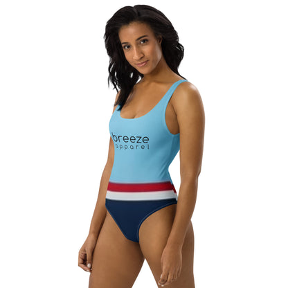 Woman wearing one-piece swimsuit with light-blue top and 'breeze apparel' in black across the chest, and a red stripe, then a white stripe, followed by a navy-blue bottom part.