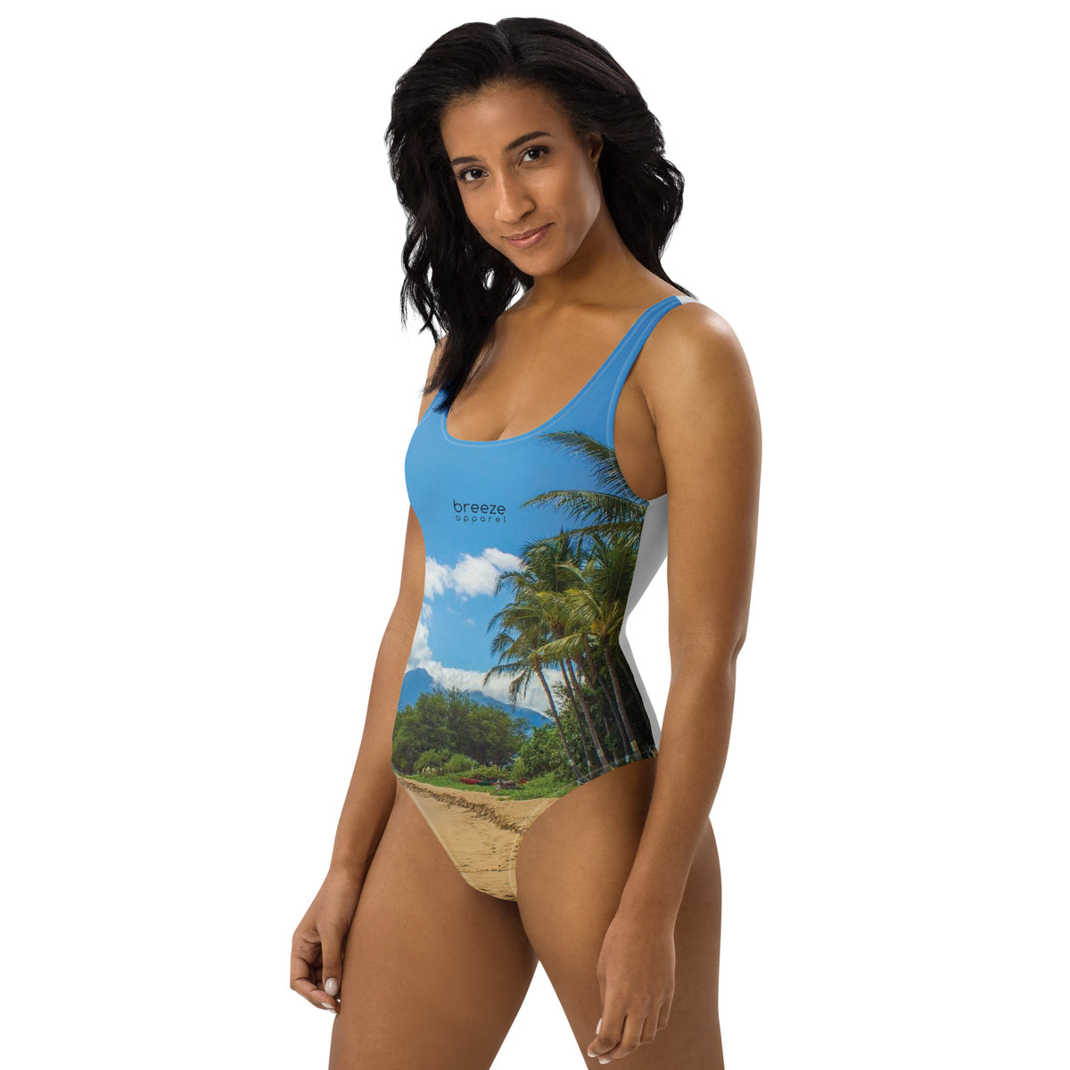 Woman wearing one-piece swimsuit with a coastline depicted on it. There is a clear sky, palm trees, mountains, and a beach depicted.