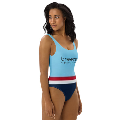 Woman wearing one-piece swimsuit with light-blue top and 'breeze apparel' in black across the chest, and a red stripe, then a white stripe, followed by a navy-blue bottom part.
