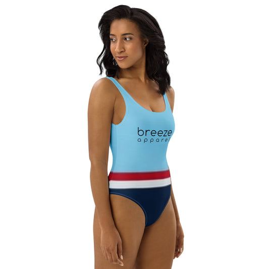 Woman wearing one-piece swimsuit with light-blue top and 'breeze apparel' in black across the chest, and a red stripe, then a white stripe, followed by a navy-blue bottom part.
