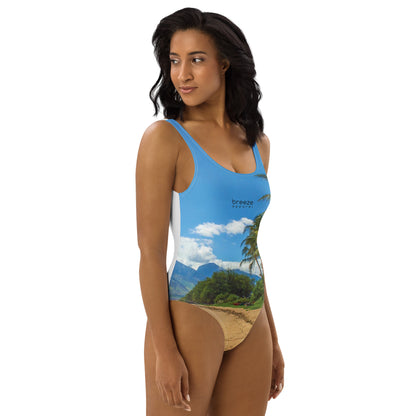 Woman wearing one-piece swimsuit with a coastline depicted on it. There is a clear sky, palm trees, mountains, and a beach depicted.