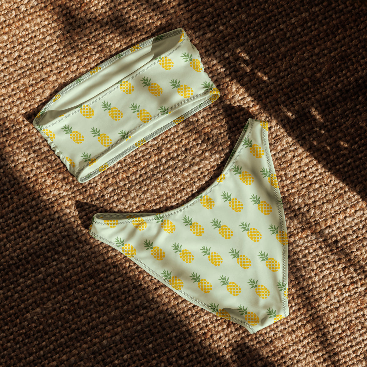 Back of bandeau bikini with an all-over pineapple design set on a light-green background, laying in the sunlight.