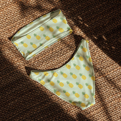 Back of bandeau bikini with an all-over pineapple design set on a light-green background, laying in the sunlight.