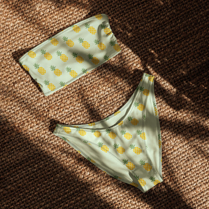 Bandeau bikini with an all-over pineapple design set on a light-green background, laying in the sunlight.