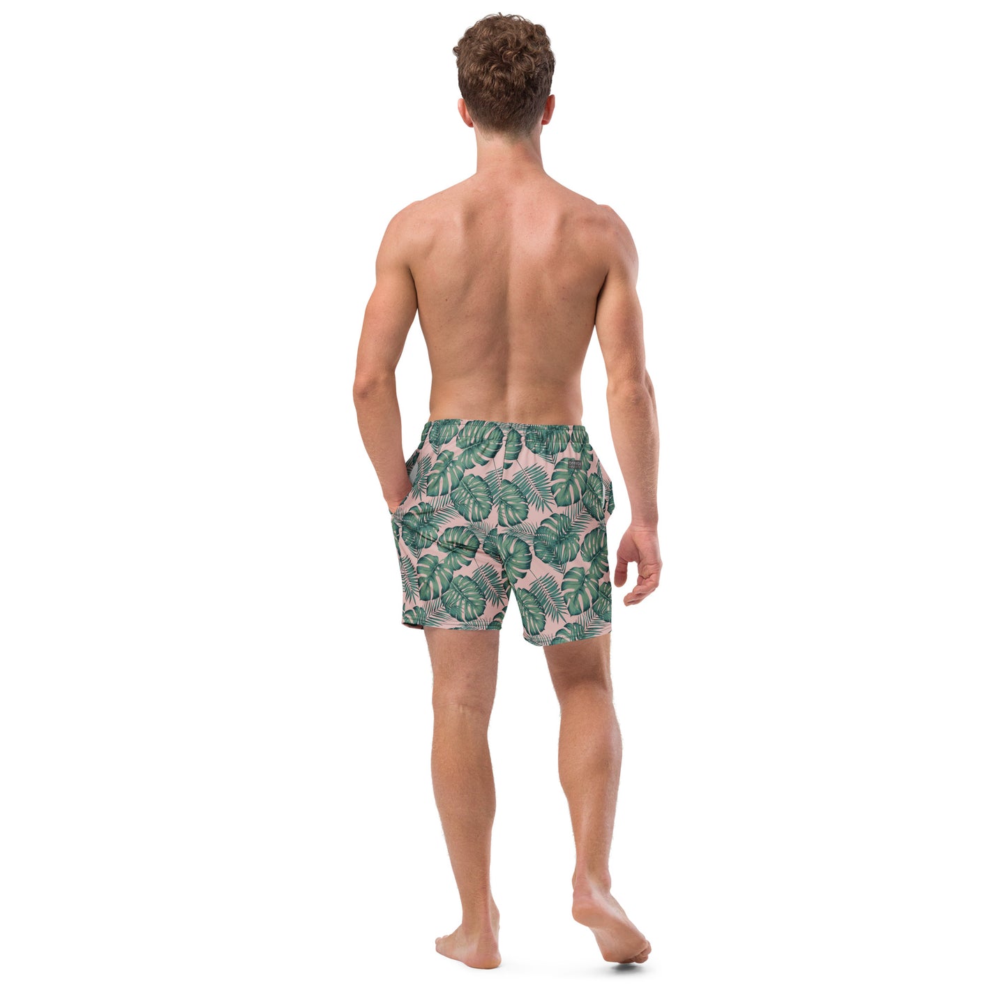 Rear view of man wearing swim trunks with pattern of green palm leaves set on a pink background.