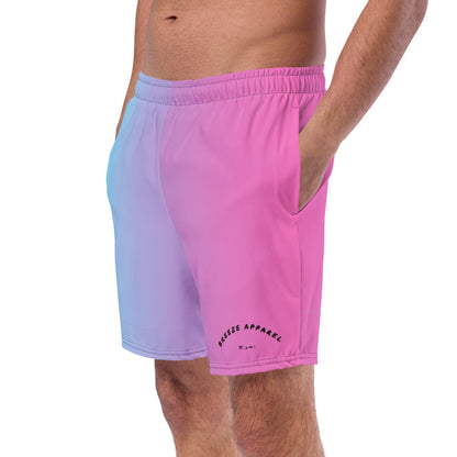 Man wearing swim trunks with a light-blue-to-pink design and a black 'Breeze Apparel Miami' word logo on the bottom-left knee