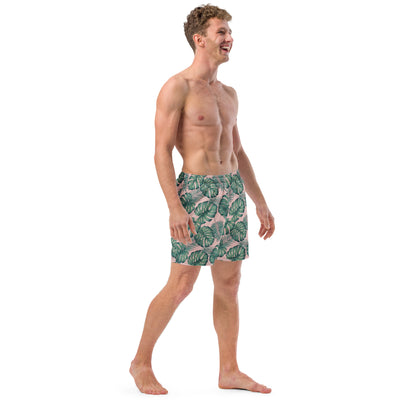 Man wearing swim trunks with pattern of green palm leaves set on a pink background.