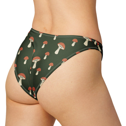 Rear view of woman wearing bikini bottom with all-over design of mushrooms set on a dark-green background, laying in the sunlight.