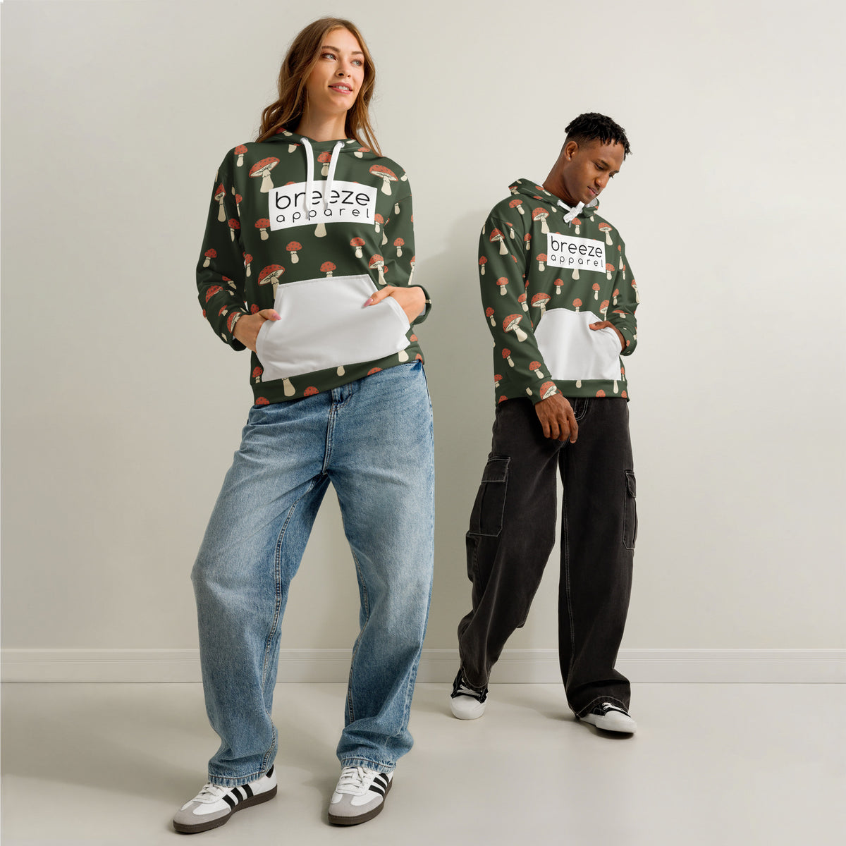 Man and woman each wearing a forest-green hoodie with all-over mushroom pattern. Hoodie interior and pocket are light gray; drawstrings are white. A white rectangle with 'breeze apparel' written in black is across the chest.