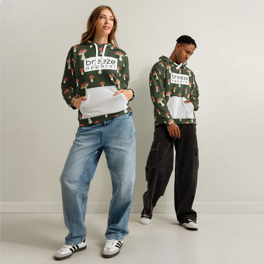 Man and woman each wearing a forest-green hoodie with all-over mushroom pattern. Hoodie interior and pocket are light gray; drawstrings are white. A white rectangle with 'breeze apparel' written in black is across the chest.