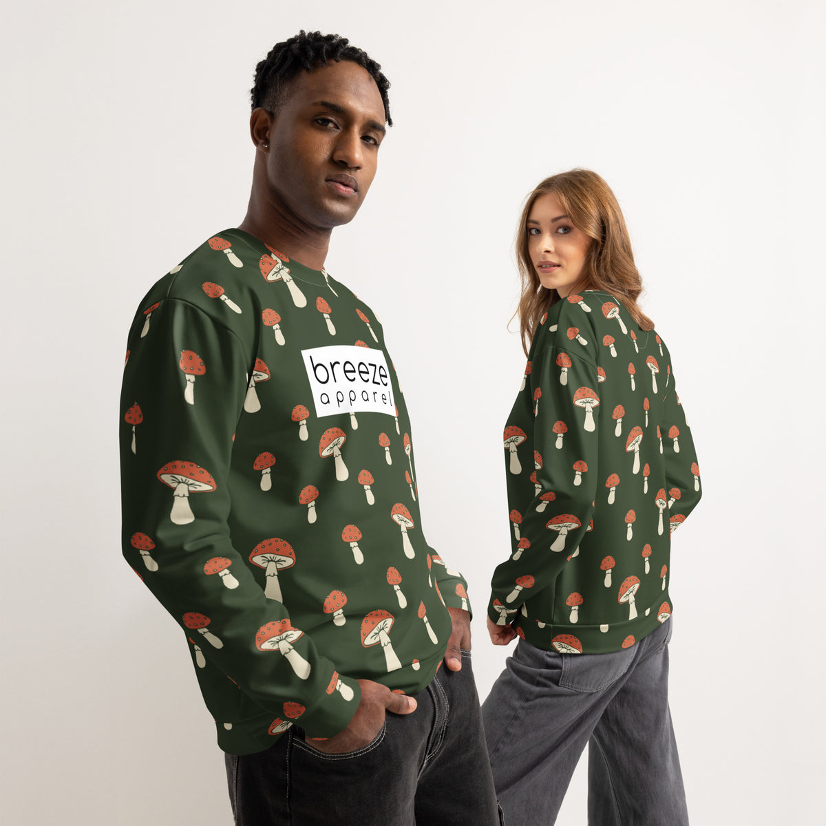 Man and woman, each wearing a forest-green sweatshirt with an all-over mushroom pattern. A white rectangle with 'breeze apparel' written in black inside is across the chest. Front is shown on man; backside is shown on woman.