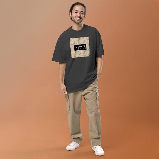 Man wearing gray, oversized t-shirt with square over chest that contains pattern of green geckos set on beige background, overlaid with black, rectangular logo with white lettering.