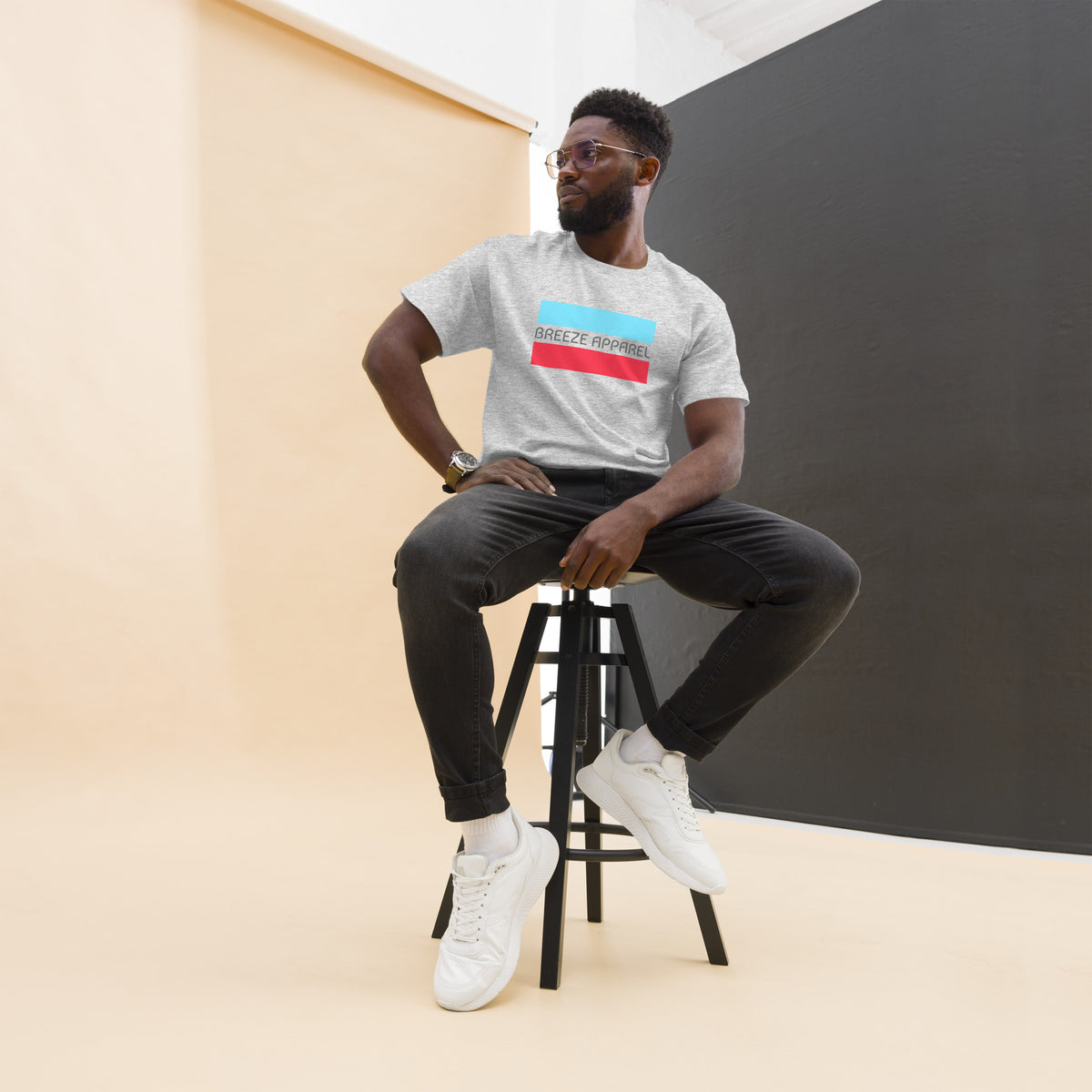 Man sitting on stool, wearing ash-gray t-shirt with gray 'Breeze Apparel' logo between horizontal bars, the top being light blue and the bottom being red