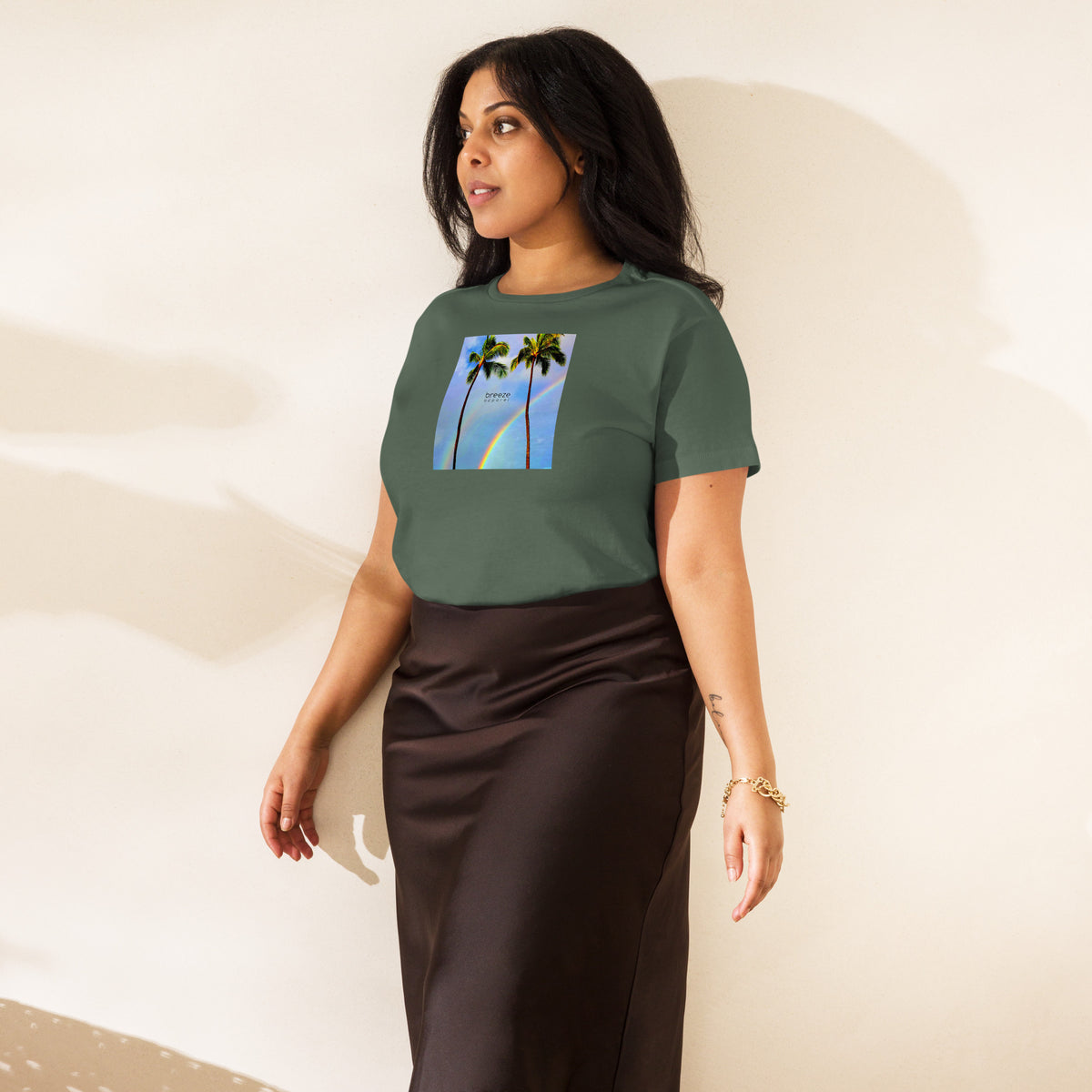 Woman wearing pine-green t-shirt with a graphic of palm trees, rainbow, and a clear blue sky in the background. A small, black 'breeze apparel' logo is between the trees.